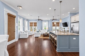 A spacious living room with a large white sofa, a kitchen with a granite countertop, and a ceiling fan.