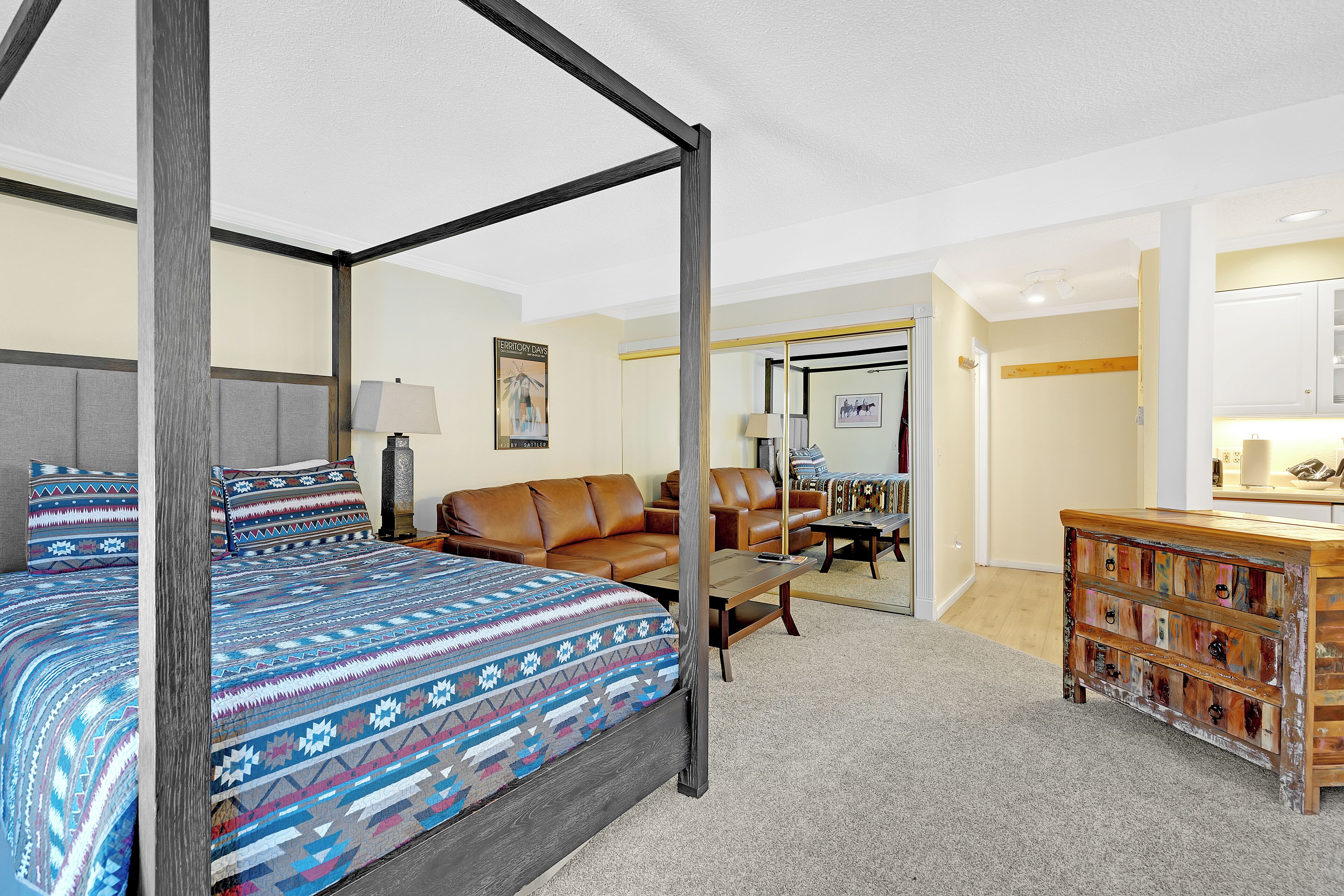 A four-poster bed with a patterned bedspread, a brown leather sofa, a wooden chest of drawers, and a small desk with a lamp.