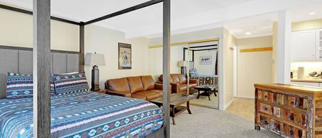 A four-poster bed with a patterned bedspread, a brown leather sofa, a wooden chest of drawers, and a small desk with a lamp.