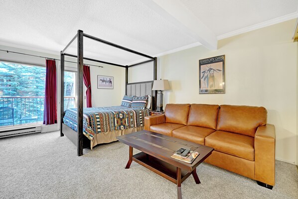 A four-poster bed with a patterned bedspread, a brown leather sofa, a wooden coffee table, and a view of a balcony with red curtains. - This Location Can’t Be Beat! Cozy Studio in Downtown Breckenridge l DS109 (Breckenridge)