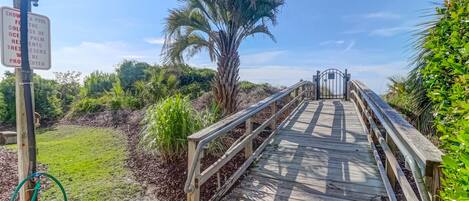 A wooden boardwalk leading to a gate, surrounded by greenery and a palm tree.