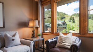 A cozy room with a view of a mountain lodge, a grey sofa, a leather armchair, and a small table with a lamp and decorative items.