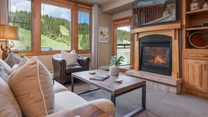 A cozy living room with a fireplace, large windows overlooking a forest, a leather sofa, and a coffee table.