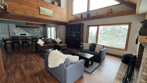 A spacious living area with a high ceiling, wooden beams, a fireplace, and large windows offering a view of trees.