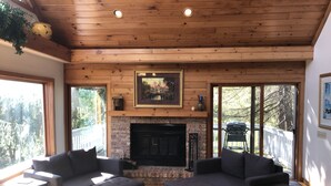 A cozy living room with a fireplace, wooden ceiling, and large windows.