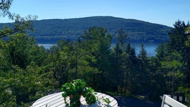 A deck with a round table and white chairs, overlooking a lake and forested hills.