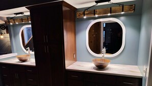 A modern bathroom with two sinks, each with a round mirror and a wooden bowl sink. The room has dark wooden cabinets and a light-colored countertop.