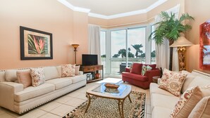 A living room with a sofa, armchairs, a glass coffee table, a flat-screen TV, and a large window with a view of palm trees. - Lush lobby-level condo w/ private patio (Destin)