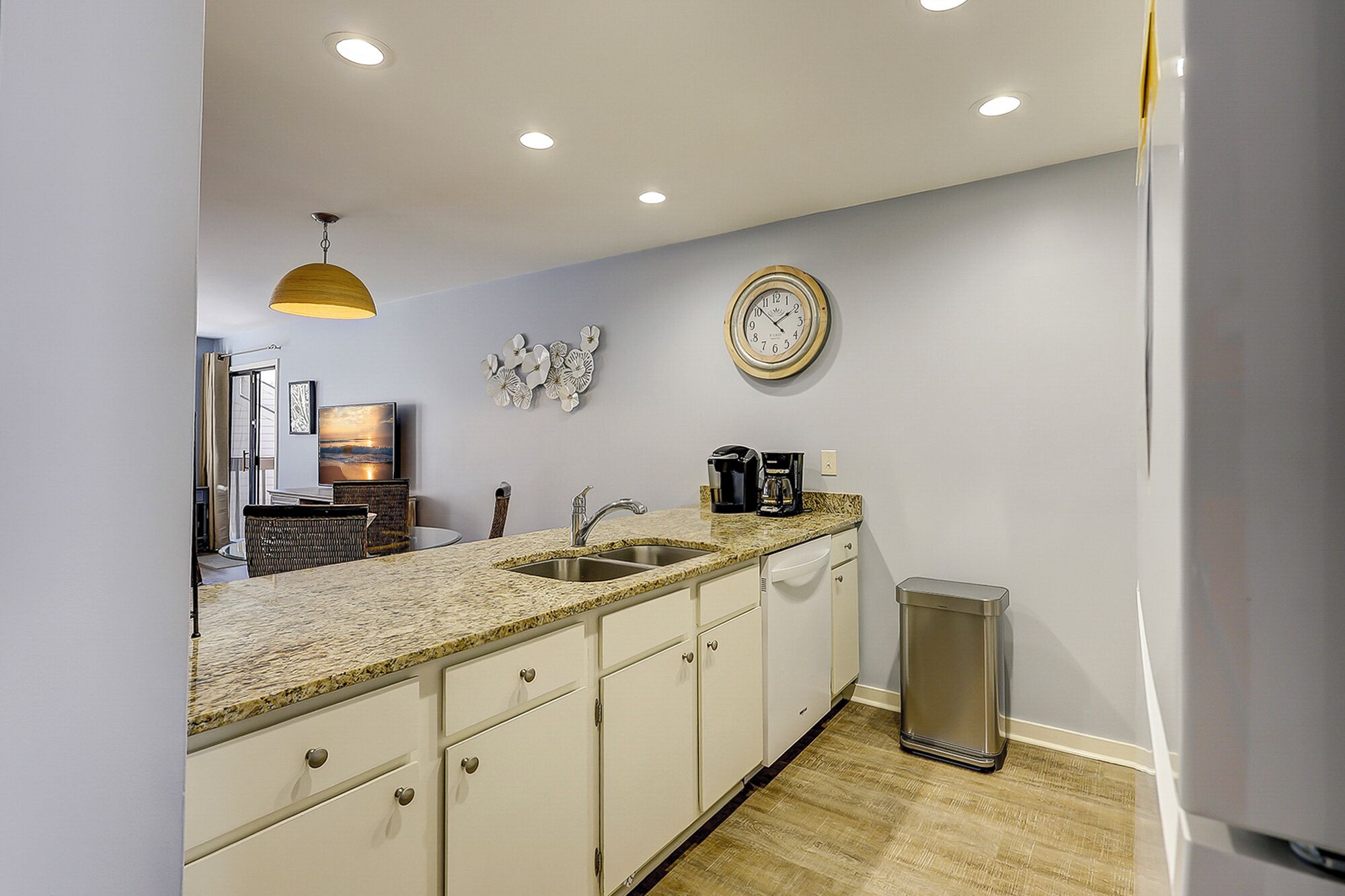 A modern kitchen with a granite countertop, white cabinetry, a stainless steel sink, a coffee maker, and a clock on the wall.