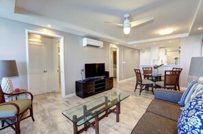 A living room with a television, a glass coffee table, a sofa, and a dining area with chairs.