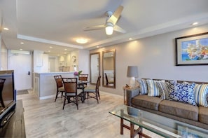 A modern living room with a glass coffee table, a sofa with patterned cushions, a dining area with wooden chairs, and a ceiling fan. - Your Perfect Poolside Haven: Royal Mauian 116 - Oceanfront Ground Floor Luxury! (Kihei, Maui)