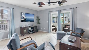 A modern living room with a large TV, comfortable seating, a ceiling fan, and a view through the glass door. - Summer Lake Haven ~ Summer Specials! ~ 2Br/2.5Ba (Miramar Beach)
