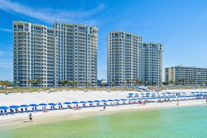 A beachfront hotel with multiple high-rise buildings, a sandy beach with blue umbrellas, and clear turquoise water.