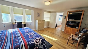 A bedroom with a bed, dresser, desk, chair, and a TV in a living area.