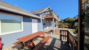 Een houten terras met picknicktafels, een overdekte patio en uitzicht op een woongebouw met een balkon.