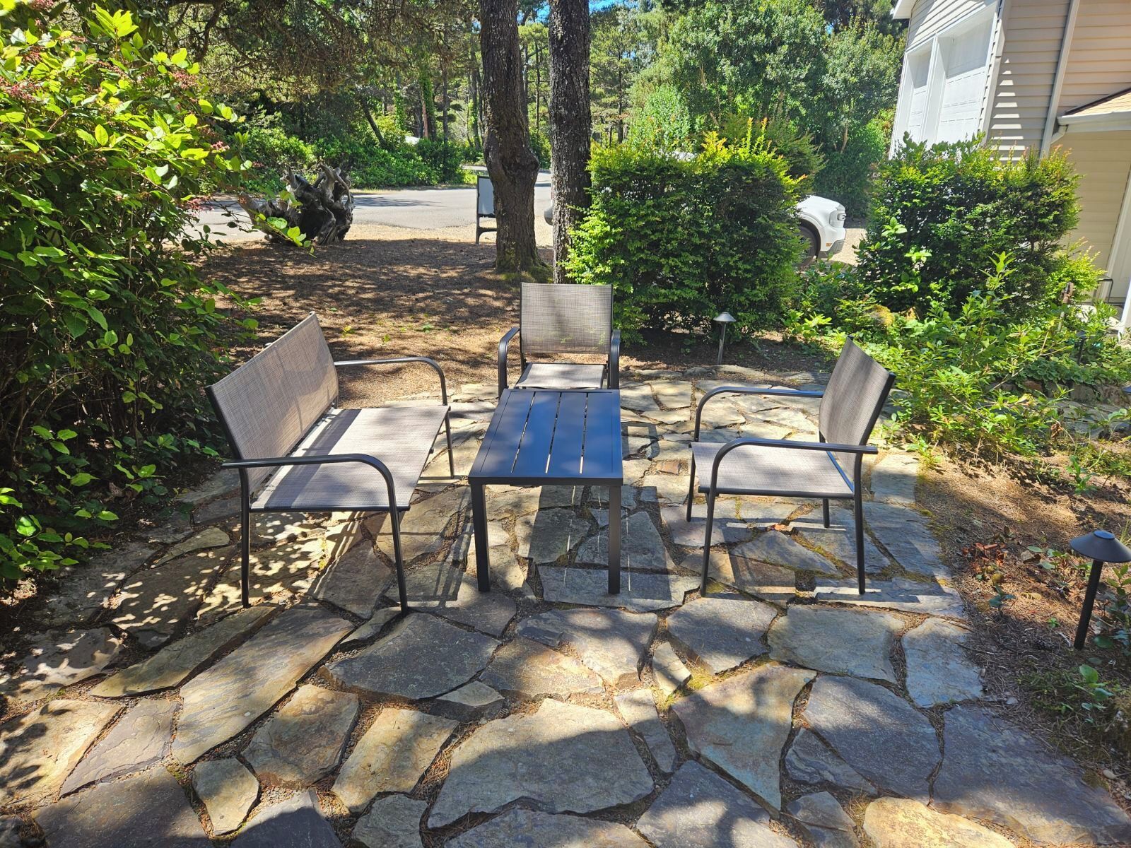 A stone patio with a table and four chairs, surrounded by greenery and trees.