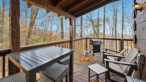 A wooden deck with a table and chairs, a grill, and a view of trees.