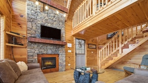 A cozy cabin interior with a stone fireplace, wooden walls, and a staircase.