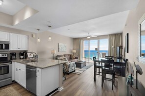 A modern kitchen with white cabinets, a granite countertop, and stainless steel appliances. There is a dining area with a table and chairs, and a balcony with a view of the ocean.