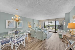 A spacious living room with a glass dining table, white chairs, a sofa, and a chandelier.