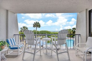 A balcony with white chairs and a table, overlooking a swimming pool and palm trees.