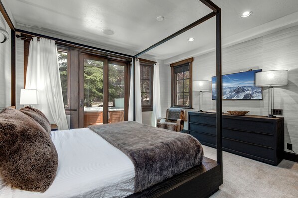 A modern bedroom with a large bed, a wooden dresser, and a view of the outdoors through large windows.