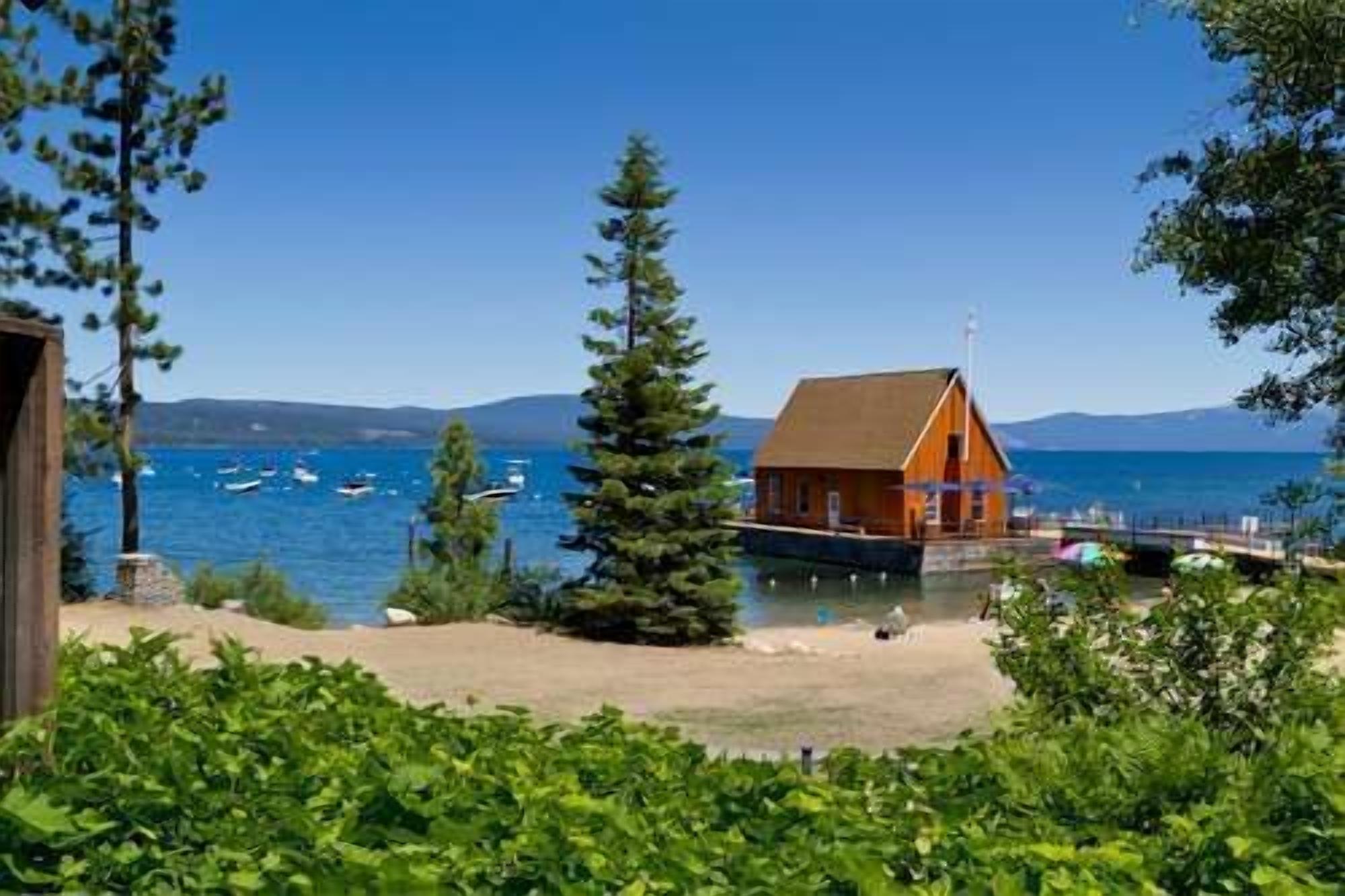 Une cabane en bois, dotée d’un toit en pente, surplombe un lac où flottent des bateaux, et est entourée de verdure, avec des montagnes qui se dessinent à l’horizon.