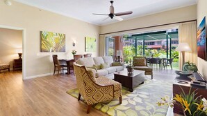 A spacious living room with a sofa, armchairs, a coffee table, and a dining area with chairs and a table. There is a ceiling fan, a large window with a view of the outside, and artwork on the walls. - Maui Resort Rentals: Honua Kai Konea 145, Upgraded Groundfloor Interior, Huge Lawn Area, Steps to Pools/Beach/BBQs! (Lahaina)