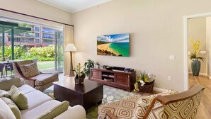 A living room with a large TV, comfortable seating, and a view of a beach through a glass door. - Maui Resort Rentals: Honua Kai Konea 145, Upgraded Groundfloor Interior, Huge Lawn Area, Steps to Pools/Beach/BBQs! (Lahaina)