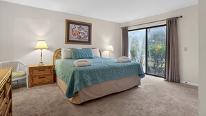 Une chambre à coucher comprenant un lit, deux tables de chevet, une chaise, une grande fenêtre avec des rideaux et un tableau encadré accroché au mur.