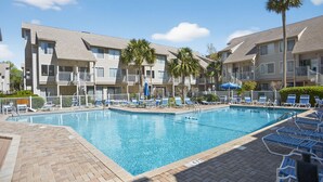 Une piscine entourée de chaises longues et de palmiers, avec des immeubles à plusieurs étages en arrière-plan.
