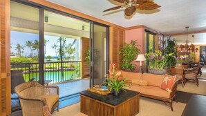 Un vaste espace de vie avec un balcon donnant sur une piscine, aménagé avec des fauteuils et un canapé en osier, ainsi qu’un ventilateur de plafond.