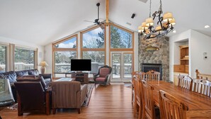 A spacious living room with a large wooden dining table, comfortable seating, a fireplace, and large windows offering a view of the outdoors.