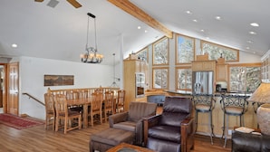 A spacious living area with a high ceiling, wooden dining table, leather sofas, and a kitchen with large windows.
