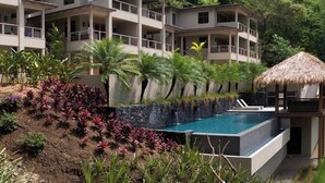 Et hotel i flere etager med swimmingpool, omgivet af tropiske planter og en struktur med stråtækte tage.