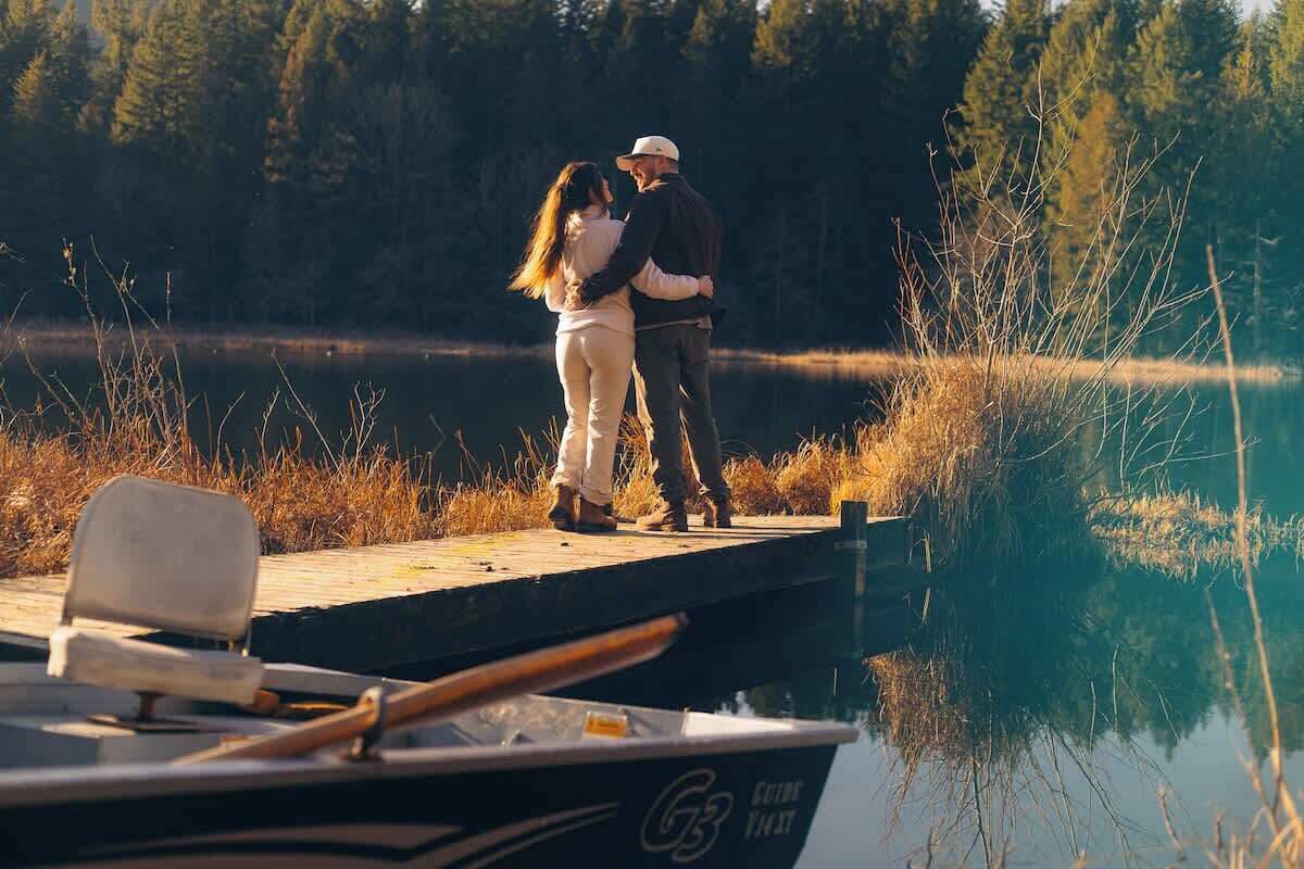 A couple standing on a dock by a calm lake, with a boat in the foreground.