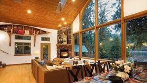 Un intérieur chaleureux avec un coin repas, un plafond en bois, une cheminée en pierre et de grandes fenêtres offrant une vue sur les arbres.