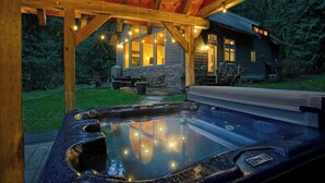 Un bain à remous sous une pergola en bois, agrémentée de guirlandes lumineuses, avec un mur en pierre et une maison en arrière-plan.