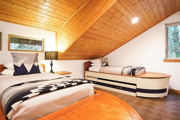 A bedroom with a sloped ceiling, two beds, a bedside table, a lamp, and a window.