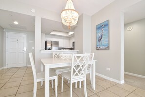 A dining area with a white table and chairs, a chandelier, and a painting of a bird on the wall. - Beachfront Condo with Views from Both Bedrooms. Includes Beach Service (Destin)