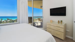 Ein Schlafzimmer mit einem großen Bett, einem an der Wand montierten Fernseher, einem Schminktisch und Blick auf den Ozean durch eine Schiebetür.