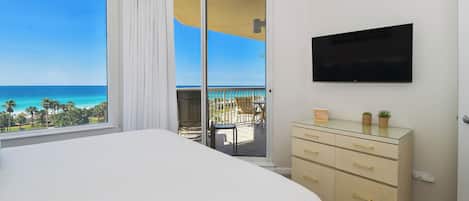 A bedroom with a large bed, a TV mounted on the wall, a dresser, and a view of the ocean through a sliding glass door.