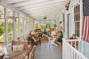 A covered porch with wooden furniture, a table, and a view of greenery. - Cozy, rustic home on Sullivan's Island near beach; great screen porch! (Sullivan's Island)