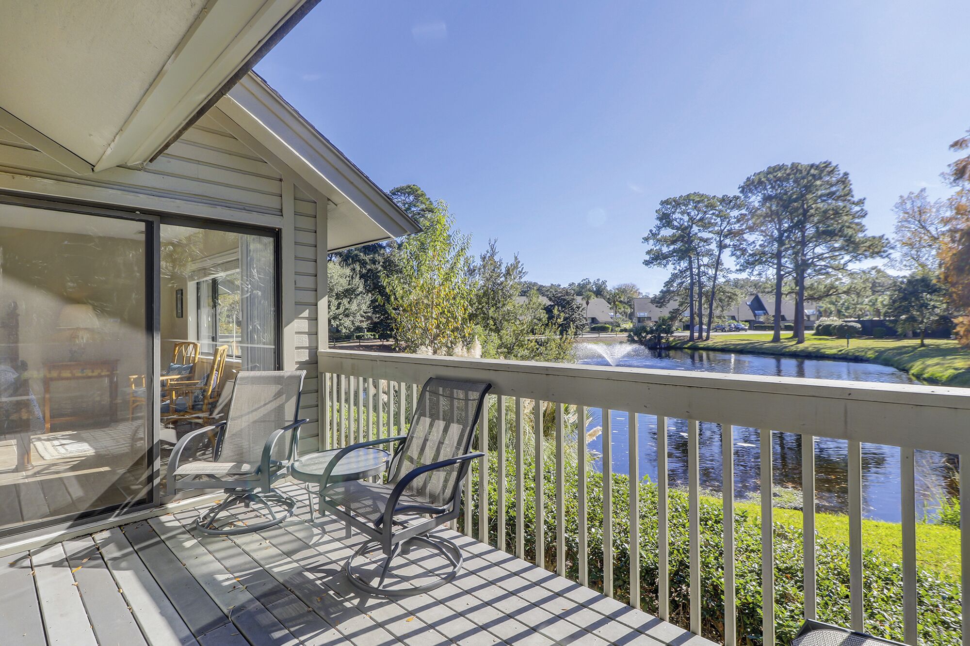 A covered outdoor deck with chairs and a table, overlooking a body of water and a landscaped area with trees and shrubs.