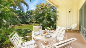 A covered outdoor seating area with a round table and white chairs, surrounded by lush greenery and palm trees. - Manualoha 603 Ground Floor By Suite Paradise (Koloa)