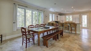 Un'ampia zona pranzo con un lungo tavolo di legno e sedie, grandi finestre con tende, e un angolo cucina con mobili bianchi e un piano di lavoro.