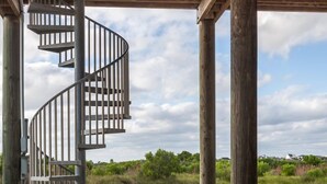 Una escalera de caracol bajo una estructura de madera, con suelo de hormigón y vistas a un campo cubierto de césped y árboles.