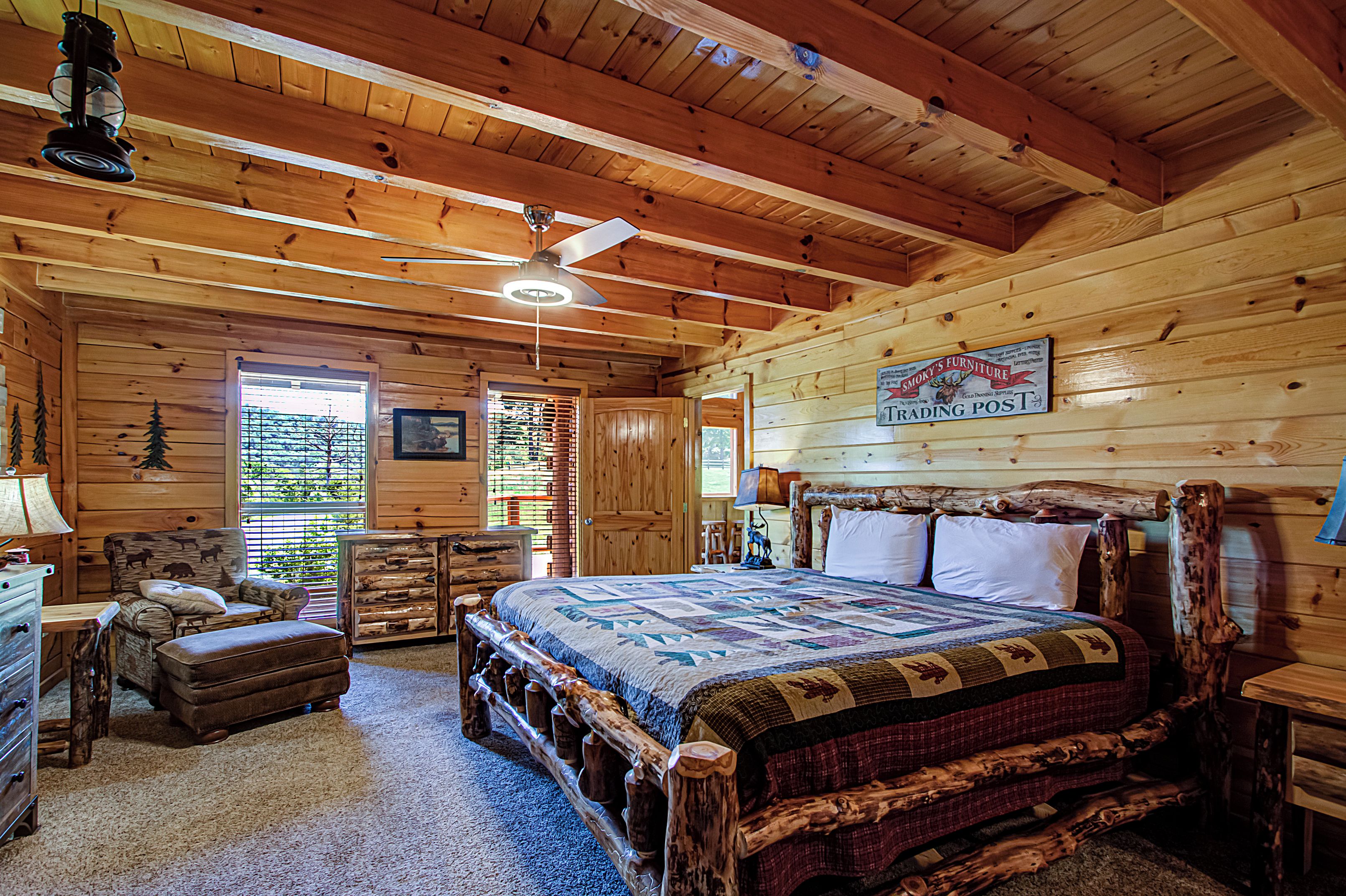 A log cabin bedroom with a wooden bed, a ceiling fan, and a sign for 'Schnitzel's Furniture Trading Post'.