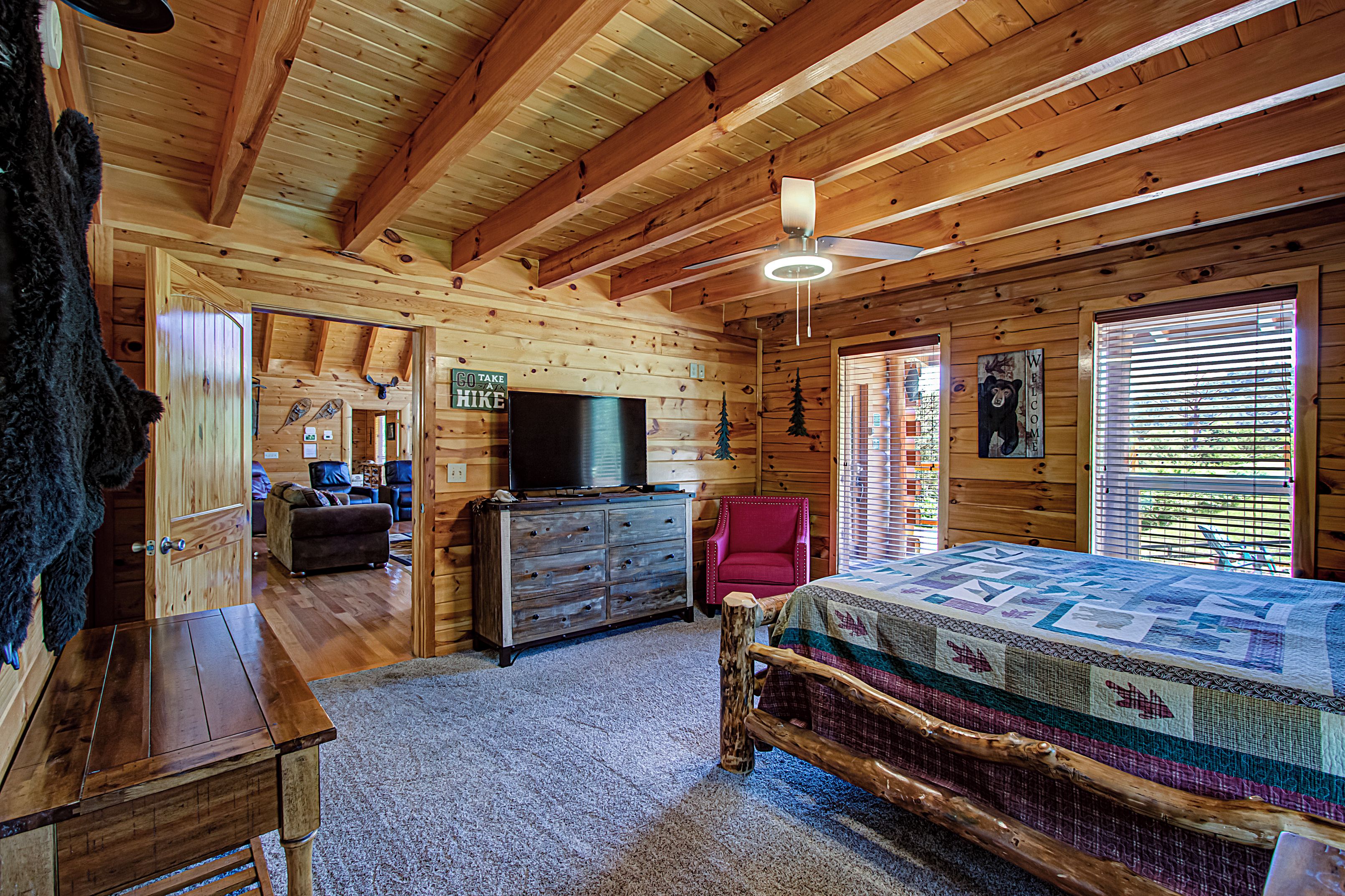 A log cabin interior with a wooden ceiling, a bed with a quilt, a television on a dresser, and a sign that says "GO TAKE HIKES".