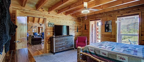A log cabin interior with a wooden ceiling, a bed with a quilt, a television on a dresser, and a sign that says "GO TAKE HIKES".